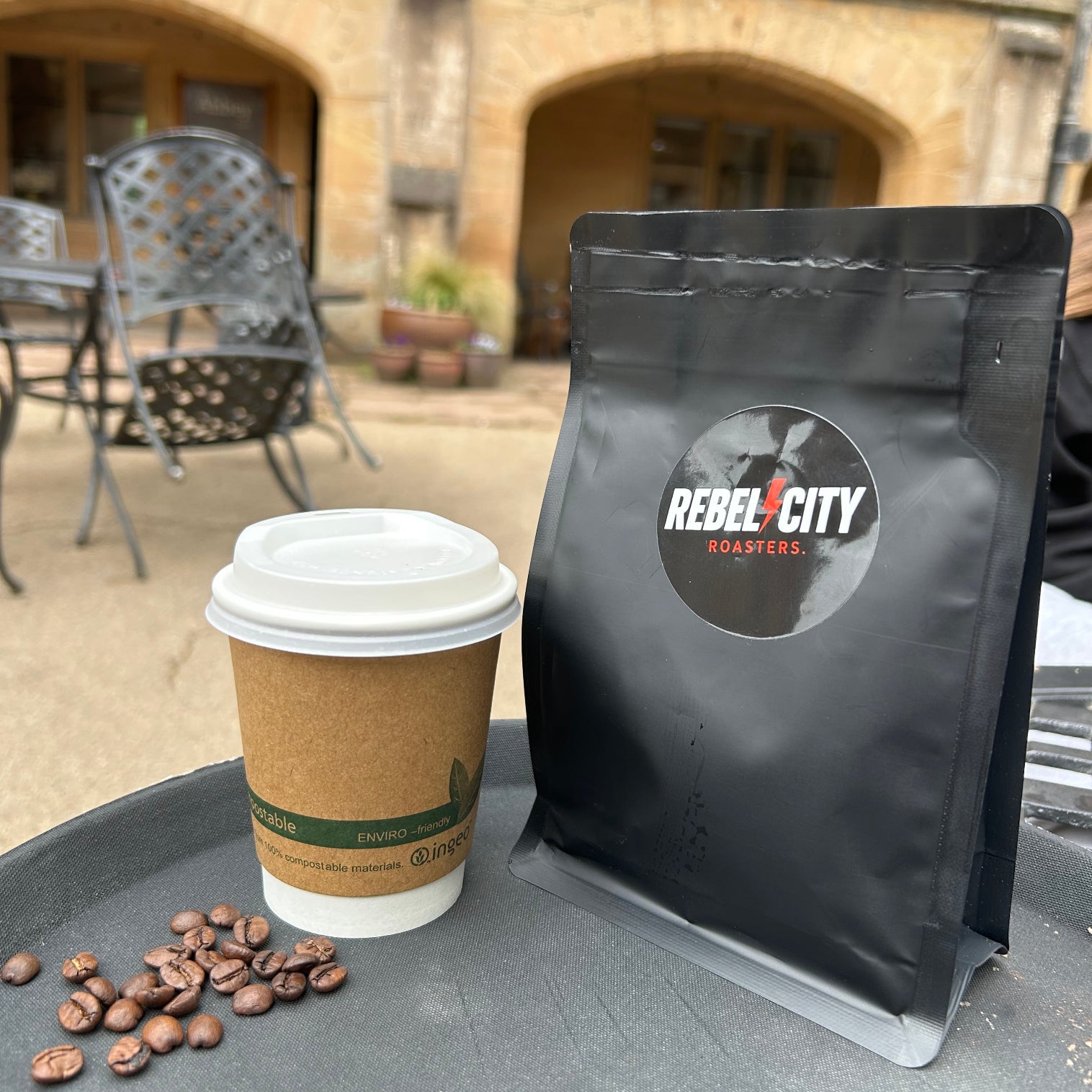 Coffee cup, coffee beans, and black bag with 'Rebel City Roasters' logo on a table outdoors.
